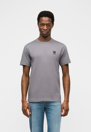 Muž v jednoduchém šedém tričku s krátkým rukávem a malým černým logem na hrudi, s světle modrými džínami, na bílém pozadí.