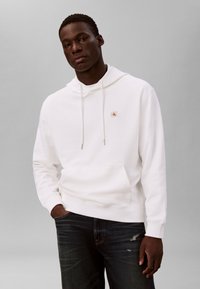 Mann iført en hvit hettegenser med en liten rød logo på brystet og mørkeblå jeans med slitte detaljer, stående med den ene hånden i lommen.