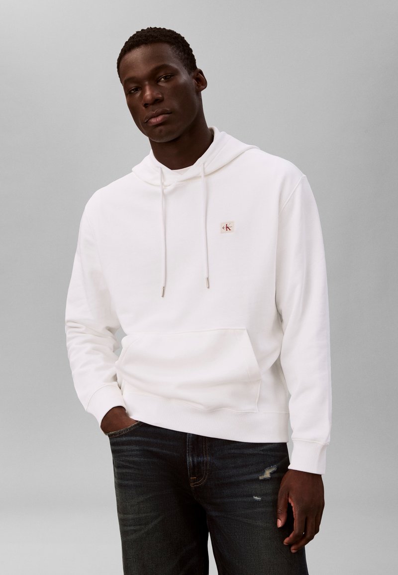 Mann iført en hvit hettegenser med en liten rød logo på brystet og mørkeblå jeans med slitte detaljer, stående med den ene hånden i lommen.