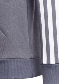 Sweatshirt gris avec un tissu texturé, doté d'un ourlet à côtes et d'une poche, agrémenté de rayures verticales blanches le long des manches.