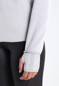 Top de manga larga gris claro con puños ajustados y un patrón texturizado, combinado con leggins negros que tienen detalles de malla.