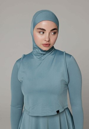 Woman wearing a fitted light blue hijab and long-sleeve top, looking to her left against a plain gray background.