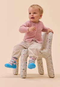 Lichtroze gebreide cardigan, crèmekleurige ribbroek en blauwe schoenen met roze banden. Zittend op een gestructureerde, gespikkelde stoel van gerecycled materiaal.