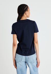 Navy blå bomulls t-skjorte med korte ermer, rund hals og løs passform, brukt med lyseblå denimshorts. Bakre utsikt viser flettehår.
