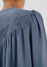 Blaue Bluse mit Puffärmlen und besticktem Blumenmuster an der Passe. Weicher Stoff mit plissiertem Design entlang der Schultern.