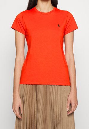 Femme portant un t-shirt orange vif à manches courtes avec un petit logo brodé, associé à une jupe plissée beige.