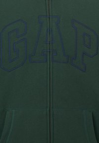 Sudadera verde con cremallera que presenta un gran logo de "GAP" en bordado azul marino, con una textura suave y bolsillos frontales.