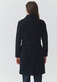Navy wool-blend coat, mid-length, with a belted waist, large collar, and smooth texture. Rear view shows clean lines and minimal detailing.