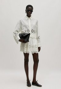 Witte katoenen shirtjurk met laser-cut kantafwerking aan de zoom en mouwen, gecombineerd met een zwarte clutch en zwarte platte schoenen.