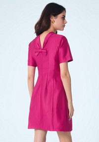 Femme portant une robe magenta à manches courtes avec un dos en V et un petit nœud, se tenant contre un fond clair simple.