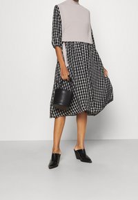 Abito a scacchi in bianco e nero con top lavorato a maglia beige. Presenta maniche a sbuffo, una gonna svasata e una borsa cilindrica nera. Mules neri.