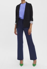 Černý blazer přes světle modrou košili, doplněný o námořnické kalhoty a zelené vysoké podpatky. Pruhovaný detail je viditelný na manžetách rukávů.