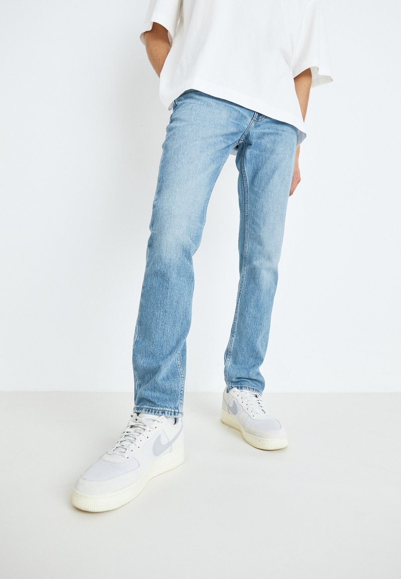 Jeans de mezclilla azul claro con un corte recto, que presentan un ligero desvanecimiento. Combinados con zapatillas gris claro y una camiseta blanca de gran tamaño.