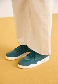 Teal värvi sneakersid, mis on valmistatud sametist ja võrgumaterjalist, valge kummitald ja silmapaistvad paelad. Lahtised kreemjad püksid, mis puutuvad kokku kingade kohal.