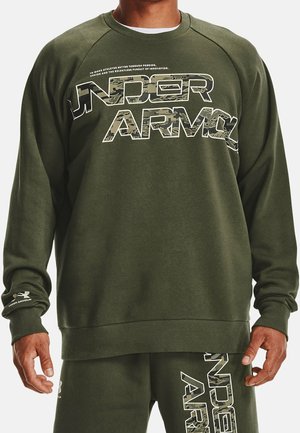 Ein Mann trägt einen olivgrünen Under Armour Sweatshirt und passende Jogginghose, beide mit großem Camouflage-Muster-Logo-Text.