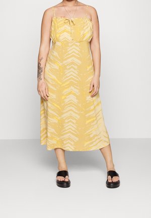 Kobieta ubrana w żółtą, wzorzystą, bezrękawnikową sukienkę midi na cienkich ramiączkach oraz czarne sandały na platformie, na jednolitym tle.