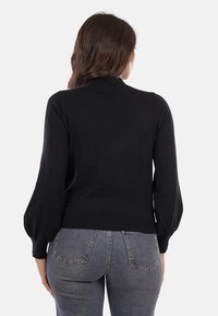 Maglione nero lavorato a maglia con maniche lunghe e puffate e collo alto. Presenta un orlo aderente. Il modello lo indossa con jeans a vita alta.