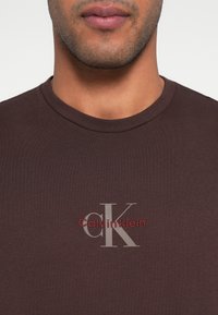 Σκούρο καφέ βαμβακερό T-shirt με στρογγυλή λαιμόκοψη. Διαθέτει γκρίζο και κόκκινο λογότυπο «Calvin Klein» και μικρές λεπτομέρειες. Ομαλή υφή.