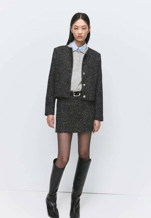 Woman wearing a black tweed jacket and matching skirt, grey polo shirt, black belt, sheer tights, and black knee-high boots against a plain background.