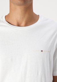 Hvit bomull T-skjorte med rund hals, subtil tekstur og en liten brodert logo i gull med røde og blå detaljer.
