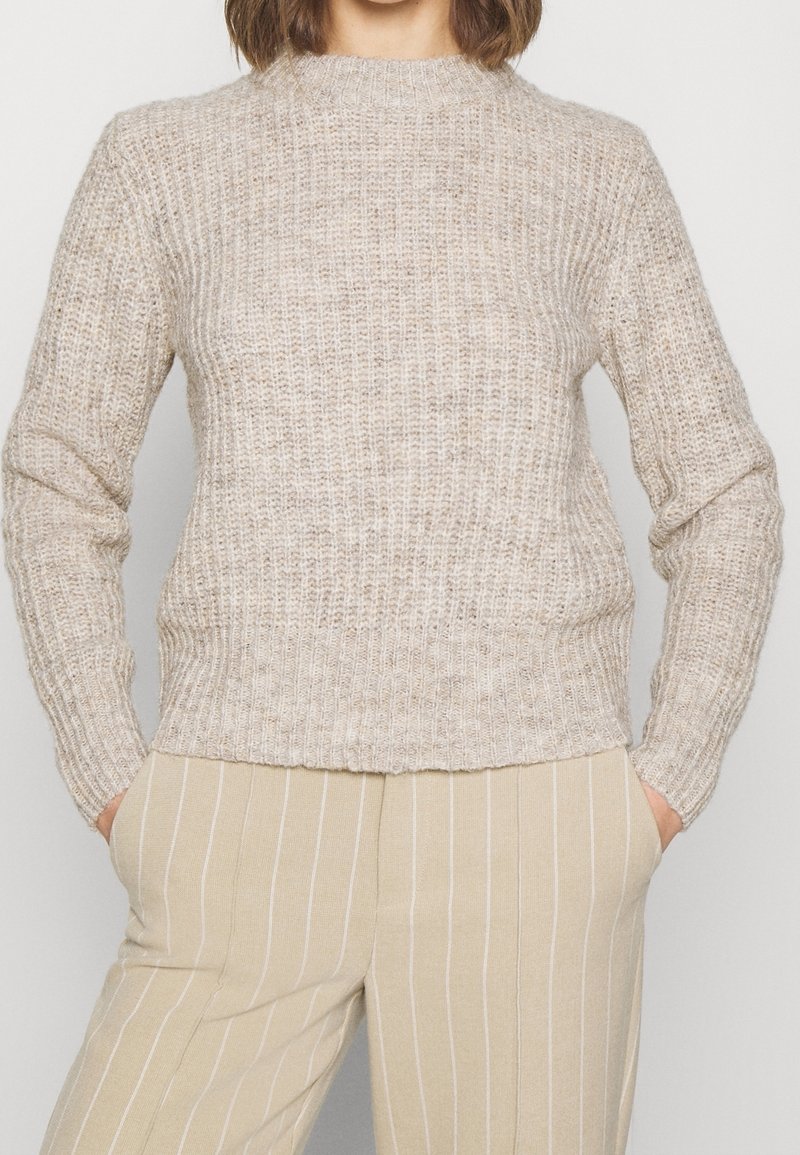 Femme portant un pull en maille côtelée beige et un pantalon beige à fines rayures, les mains dans les poches, sur un fond uni.