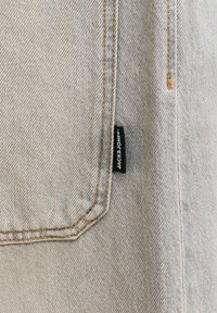 Tissu en denim gris clair avec des surpiqûres orange définissant une poche, avec une étiquette en tissu noire portant l'inscription "JACK & JONES."