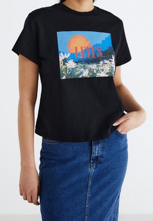 Osoba nosząca czarną koszulkę z kolorową grafiką pola kwiatów i słońca oraz czerwonym napisem "LEVI'S", połączoną z niebieską spódnicą jeansową.