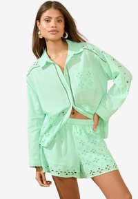 Lichtgroene katoenen set met een knoopsluiting shirt met oogjes uitsnijdingen en bijpassende shorts met bloemmotieven en een elastische tailleband.