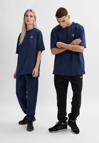 Marinblå bomulls-t-shirts och byxor som bärs av modeller, med avslappnad passform, korta ärmar och en liten logotyp. Ett par är svarta, det andra marinblå.
