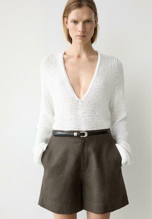Femme portant un pull en tricot blanc à col en V profond, rentré dans un short marron taille haute avec une ceinture noire, posant devant un fond uni.