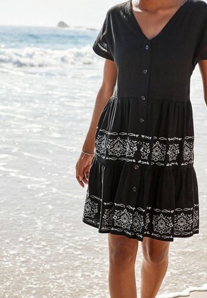 Schwarzes Hemdkleid mit V-Ausschnitt, kurzen Ärmeln, gerafften Stufen und weißer Stickerei am Saum, die florale und Fischmuster darstellt.