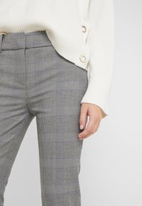 Grå rutiga byxor med ett texturerat tyg, som har svarta och blå linjer. En krämfärgad tröja med synliga knapppdetaljer i nederkant.