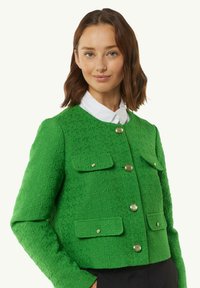 Groene gestructureerde jas met een ronde halslijn, sluiting met knopen aan de voorkant en twee klepzakken. Voorzien van gouden hardware en lange mouwen.