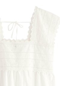 Vit klänning med fyrkantig halsringning, vågiga kanter och spetsdetaljer. Har en knytning bak och en texturerad bodice med blommönster.