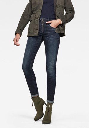 Persoon die donkerblauwe slim-fit jeans draagt met omgeslagen boorden, olijfgroene enkellaarzen met rits, een marineblauwe trui en een olijfgroene jas.