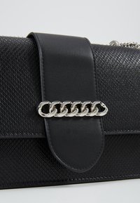 Borsa nera con superficie testurizzata e pattina in pelle liscia, fissata da un accento in catena argentata. Design compatto con dettagli sottili.