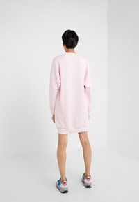 Person som står vänd bort och har på sig en lös, långärmad rosa sweatshirtklänning och färgglada sneakers i ett vitt rum.