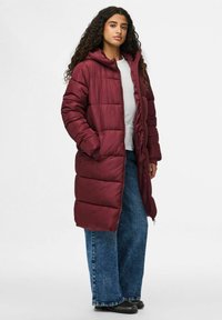 Bordo pūkinis paltas su gobtuvu, turintis horizontalius siuvinėjimus ir viso ilgio užtrauktuką. Dėvimas ant baltos marškinėlių ir mėlynų plačių džinsų.