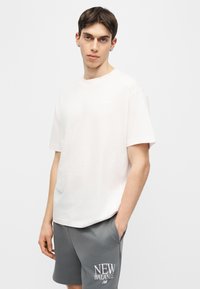 Νέος άνδρας με κοντά σκουρόχρωμα μαλλιά, που φοράει λευκό φαρδύ T-shirt και γκρι σορτς New Balance, με τα χέρια στις τσέπες, ουδέτερο φόντο.