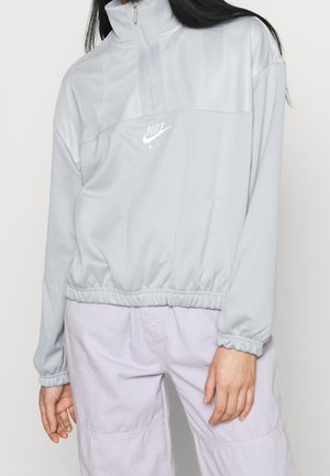 Osoba nosząca jasnoszary pulower Nike Air z półzamkiem, elastycznym ściągaczem i mankietami, w połączeniu z jasnofioletowymi spodniami, stojąca na jednolitym tle.