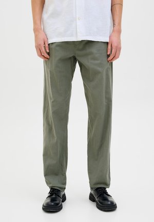 Hombre vistiendo pantalones rectos de color verde oliva, zapatos negros con cordones y una camisa blanca de botones frente a un fondo blanco.