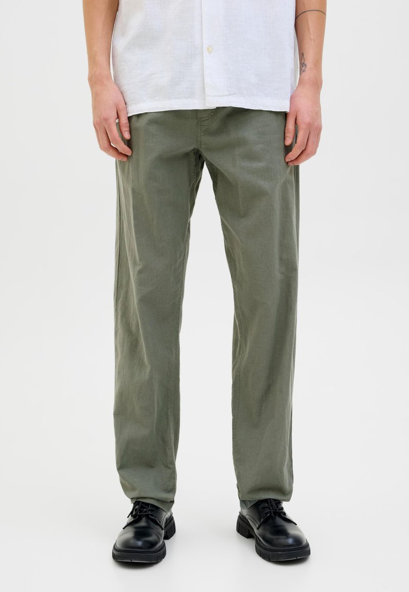 Hombre vistiendo pantalones rectos de color verde oliva, zapatos negros con cordones y una camisa blanca de botones frente a un fondo blanco.