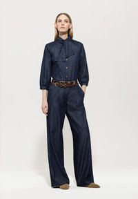 Woman wearing a dark blue jumpsuit with gold buttons, a brown belt, and tan shoes, standing against a plain light background.