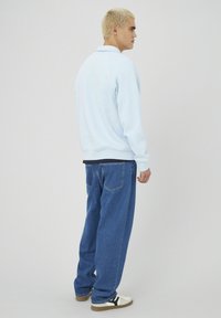 Homme à cheveux blonds courts portant un sweat-shirt bleu clair, un jean bleu ample et des baskets blanches, se tenant de profil devant un fond uni.