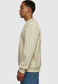 Sudadera beige con patrón de líneas negras onduladas, puños acanalados y cuello redondo. Modelo mostrado de perfil, combinado con jeans azules.