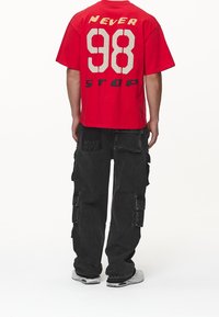 Camiseta roja de gran tamaño con la inscripción "NEVER 98 STOP" en letras blancas y negras. Combinada con pantalones cargo negros que presentan múltiples bolsillos y detalles de logo.
