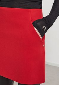 Rode stoffen rok met een zijzak, een gladde textuur en een subtiele glans. Ernaast een zwarte blouse met lange mouwen met een decoratieve accent.