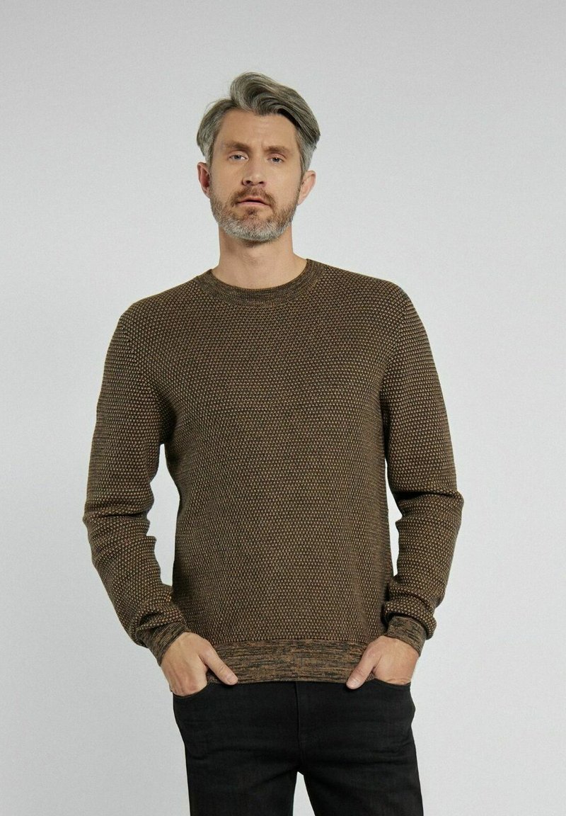 Brązowy sweter wykonany z dzianiny, z fakturowanym wzorem, okrągłym dekoltem oraz ściągaczami na rękawach i w dole. Nosi się go z czarnymi spodniami.