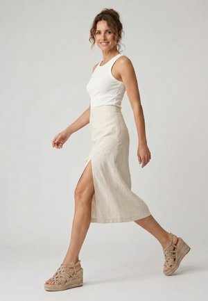 Frau geht selbstbewusst in weißem ärmellosem Oberteil, beigem Midirock mit hohem Schlitz und mit Nieten besetzten Keilsandaletten vor einfarbigem Hintergrund.