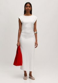 Witte, aansluitende maxidress met korte mouwen, met zijrucheing. Het model houdt een helderrode tote bag vast en draagt doorzichtige schoenen.
